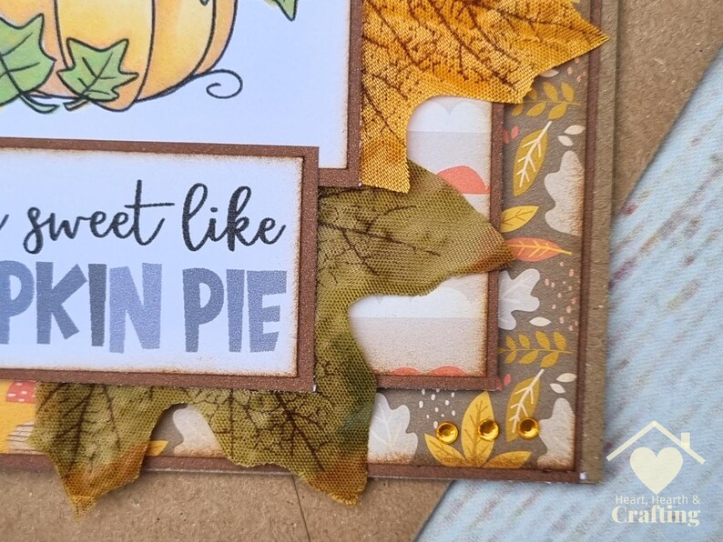 Peut inclure: Une carte de v&oelig;ux artisanale avec un design superpos&eacute;, une illustration de citrouille et le texte "sweet like PUMPKIN PIE". La carte est d&eacute;cor&eacute;e de feuilles d'automne en tissu dans des tons orange et marron, de papier &agrave; motifs et de trois petites gemmes adh&eacute;sives ambr&eacute;es.