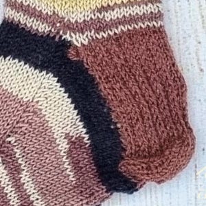 May include: A hand-knitted ankle sock featuring a striped pattern in shades of brown, cream, and black. The sock has a ribbed cuff and a reinforced heel section, showcasing a textured knit construction with distinct colour-blocked horizontal stripes.