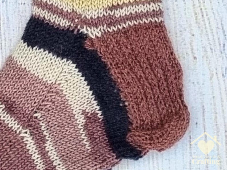 May include: A hand-knitted ankle sock featuring a striped pattern in shades of brown, cream, and black. The sock has a ribbed cuff and a reinforced heel section, showcasing a textured knit construction with distinct colour-blocked horizontal stripes.