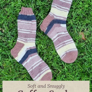 May include: A pair of hand-knitted wool socks featuring horizontal stripes in shades of mauve, cream, and dark navy blue. The text on the image reads: Soft and Snuggly Coffee Socks, Handknitted with Wool, www.hearthearthcrafts.com.