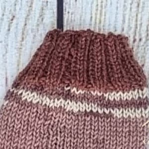 May include: A hand-knitted brown sock featuring a ribbed cuff and a single horizontal cream-coloured stripe. The knit texture is visible throughout the garment, which is displayed against a light, textured background.