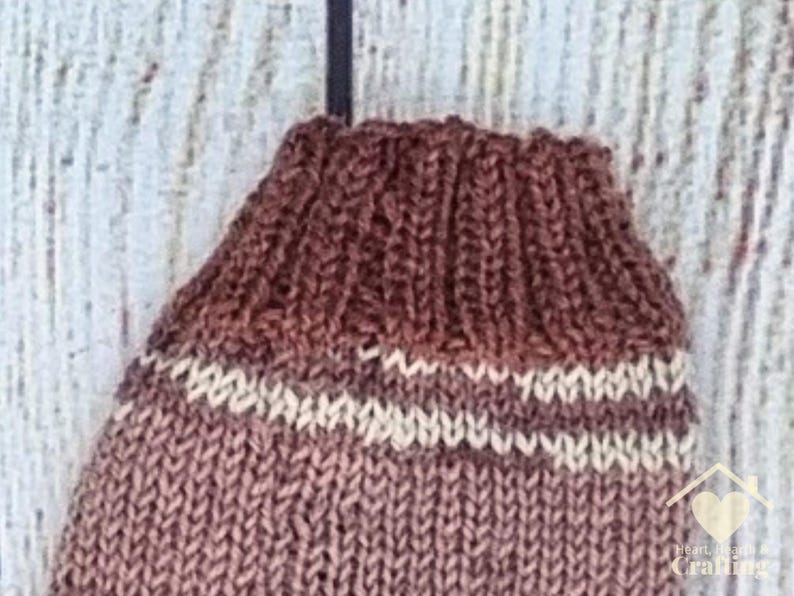 May include: A hand-knitted brown sock featuring a ribbed cuff and a single horizontal cream-coloured stripe. The knit texture is visible throughout the garment, which is displayed against a light, textured background.
