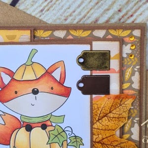 Peut inclure: Une carte de v&oelig;ux artisanale repr&eacute;sentant un renard de dessin anim&eacute; portant un chapeau citrouille et assis sur une citrouille. La carte est orn&eacute;e de deux petites &eacute;tiquettes m&eacute;talliques, d'une feuille d'automne en tissu et de strass jaunes.