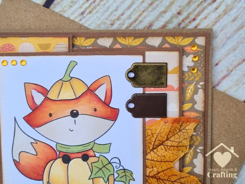 Peut inclure: Une carte de v&oelig;ux artisanale repr&eacute;sentant un renard de dessin anim&eacute; portant un chapeau citrouille et assis sur une citrouille. La carte est orn&eacute;e de deux petites &eacute;tiquettes m&eacute;talliques, d'une feuille d'automne en tissu et de strass jaunes.