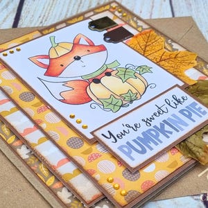 Peut inclure: Une carte de v&oelig;ux artisanale repr&eacute;sentant un renard en dessin anim&eacute; portant un chapeau citrouille et assis dans une citrouille, orn&eacute;e de feuilles d'automne et d'accents dor&eacute;s. Le texte You're sweet like PUMPKIN PIE est imprim&eacute; en bleu et noir.