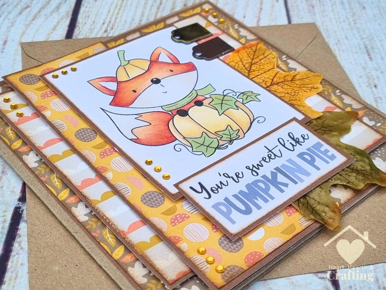 Peut inclure: Une carte de v&oelig;ux artisanale repr&eacute;sentant un renard en dessin anim&eacute; portant un chapeau citrouille et assis dans une citrouille, orn&eacute;e de feuilles d'automne et d'accents dor&eacute;s. Le texte You're sweet like PUMPKIN PIE est imprim&eacute; en bleu et noir.