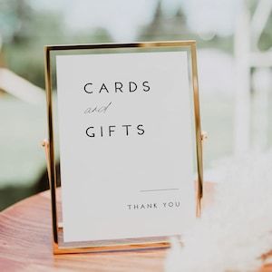 May include: A gold-framed sign with white background and black text that reads "CARDS and GIFTS" with a line and "THANK YOU" below. The sign is on a wooden surface.