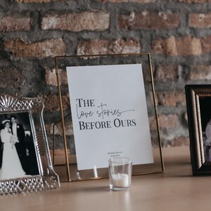 May include: A framed print with the text "The love stories BEFORE OURS" sits on a wooden surface, next to a small candle and framed photos. The frame is gold and the background is a brick wall.