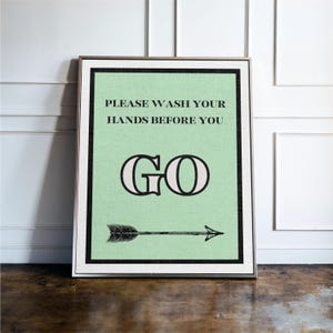 May include: Framed wall art with a mint green background and black border. The text "PLEASE WASH YOUR HANDS BEFORE YOU GO" is displayed in black, with a decorative "GO" and an arrow pointing right.