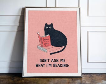 May include: A framed wall art print on a pink background featuring a black cat sitting on a stack of books. The cat holds a red book titled Enemies to Lovers. Below the cat, the text reads Don't Ask Me What I'm Reading in black capital letters.