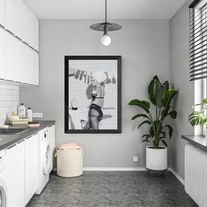 Puede incluir: Un lavadero moderno con armarios blancos, una encimera oscura y un cuadro enmarcado en negro. La habitaci&oacute;n cuenta con una lavadora, un fregadero y una gran planta en maceta. Una ventana con persianas proporciona luz natural.