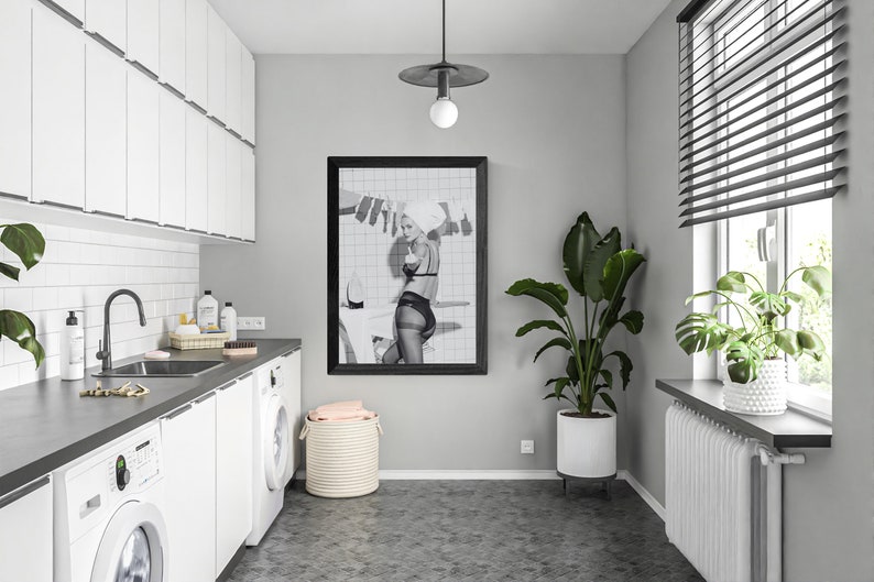 Puede incluir: Un lavadero moderno con armarios blancos, una encimera oscura y un cuadro enmarcado en negro. La habitaci&oacute;n cuenta con una lavadora, un fregadero y una gran planta en maceta. Una ventana con persianas proporciona luz natural.
