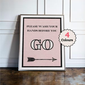 May include: Framed wall art with a pink background and black text that reads "PLEASE WASH YOUR HANDS BEFORE YOU GO" with an arrow pointing right. A circular tag indicates "4 Colours". The frame is silver.