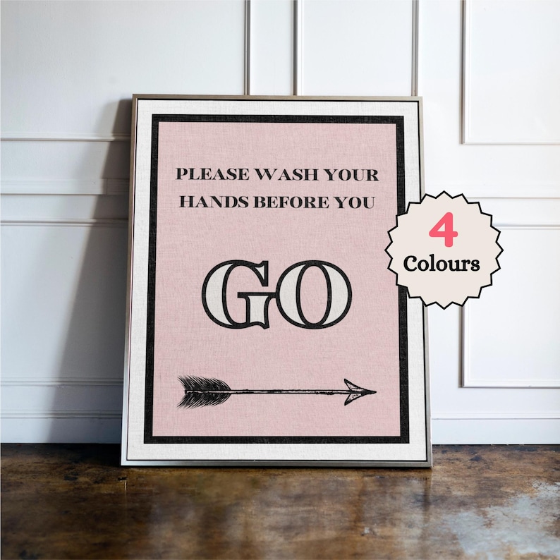 May include: Framed wall art with a pink background and black text that reads "PLEASE WASH YOUR HANDS BEFORE YOU GO" with an arrow pointing right. A circular tag indicates "4 Colours". The frame is silver.