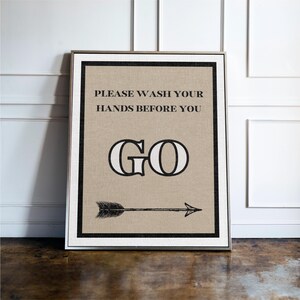 May include: Framed wall art with the message "PLEASE WASH YOUR HANDS BEFORE YOU GO." The text is in black on a tan background, with a black border and a silver frame. An arrow points to the right.