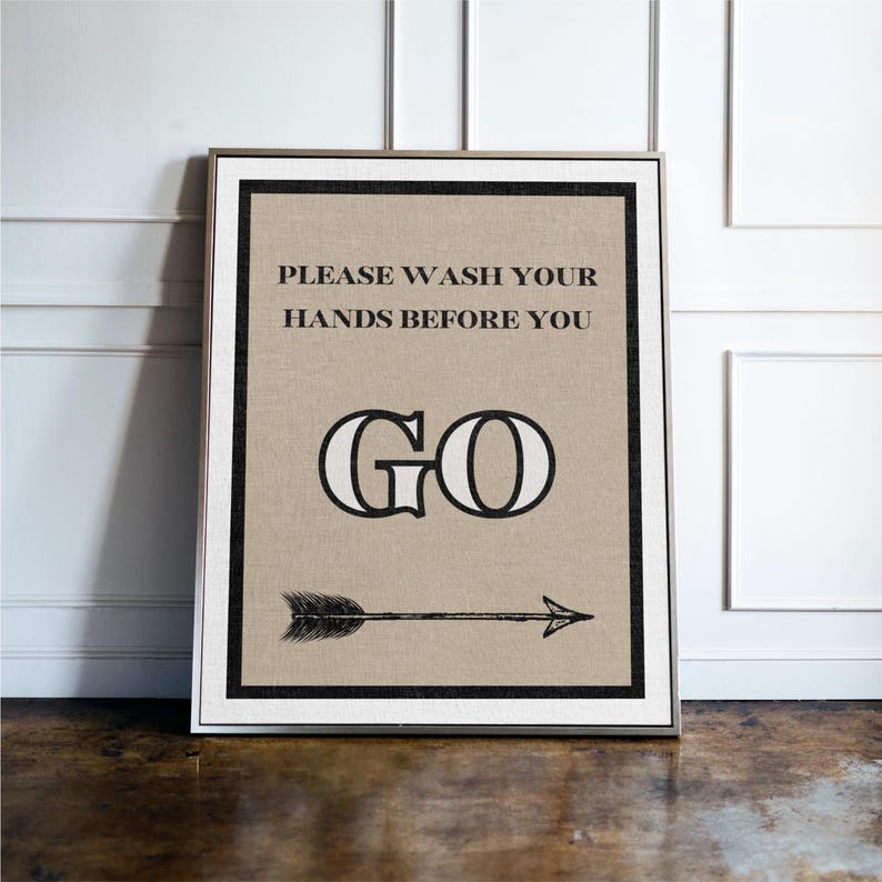 May include: Framed wall art with the message "PLEASE WASH YOUR HANDS BEFORE YOU GO." The text is in black on a tan background, with a black border and a silver frame. An arrow points to the right.