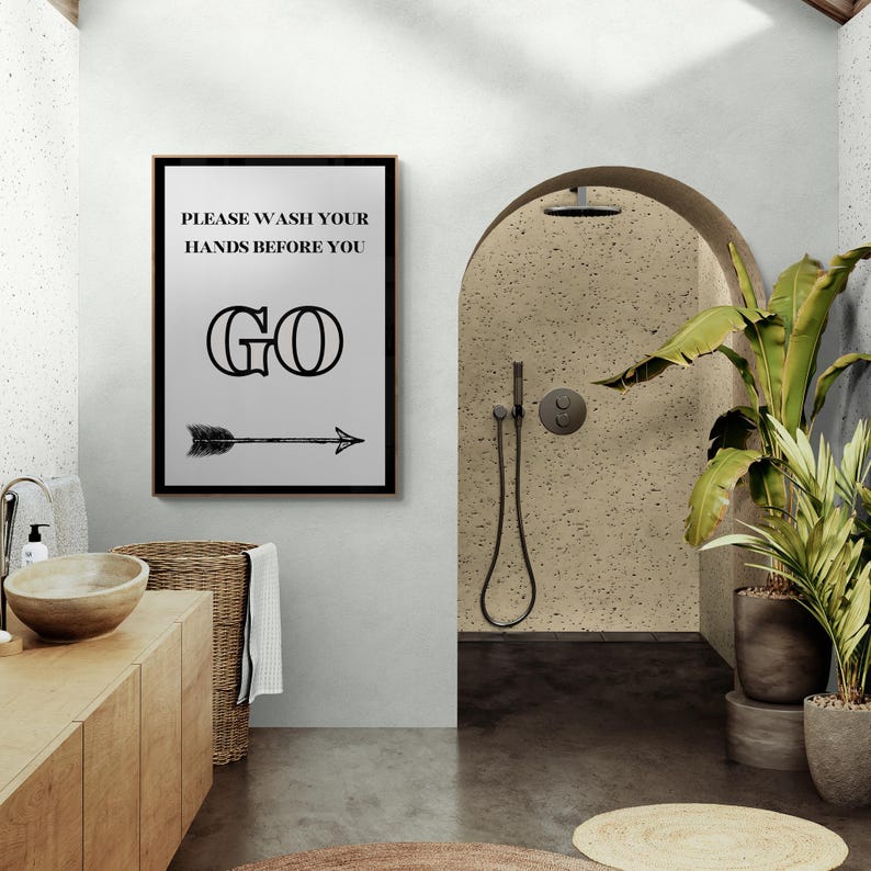 May include: A framed bathroom sign with the text "PLEASE WASH YOUR HANDS BEFORE YOU GO" in black. Below the text is a black arrow pointing right. The sign is in a modern bathroom with a wooden vanity and a shower.