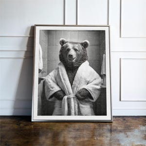 May include: A black and white photograph of a bear wearing a white bathrobe. The bear is standing in a bathroom with a towel rack and a mirror in the background.