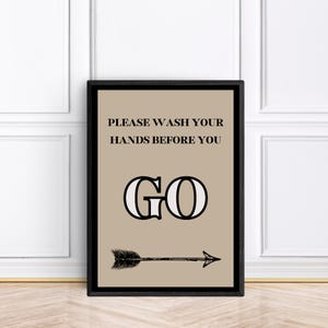 May include: A framed print with a tan background and black text that reads "PLEASE WASH YOUR HANDS BEFORE YOU GO." Below the text is a large "GO" and a black arrow pointing right. The frame is black.