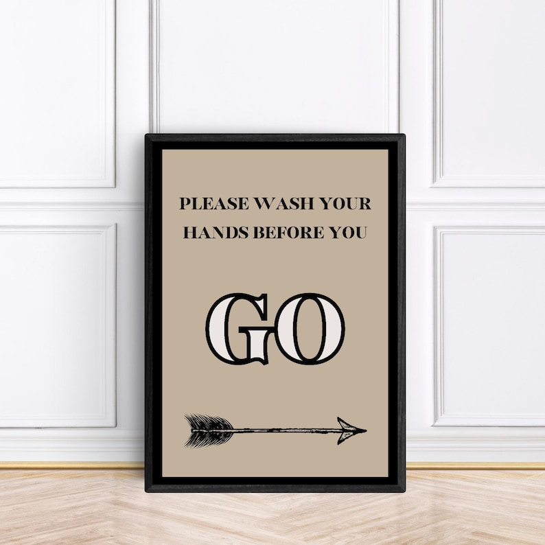 May include: A framed print with a tan background and black text that reads "PLEASE WASH YOUR HANDS BEFORE YOU GO." Below the text is a large "GO" and a black arrow pointing right. The frame is black.