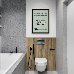 May include: A modern bathroom with a white toilet, wooden paneling, and gray tile walls. A framed sign reads "PLEASE WASH YOUR HANDS BEFORE YOU GO" with an arrow. Black fixtures and a bathtub are also visible.