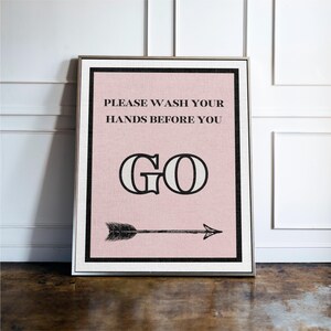 May include: Framed wall art with a pink background and black text that reads "PLEASE WASH YOUR HANDS BEFORE YOU GO." A black arrow points to the right. The frame is silver.