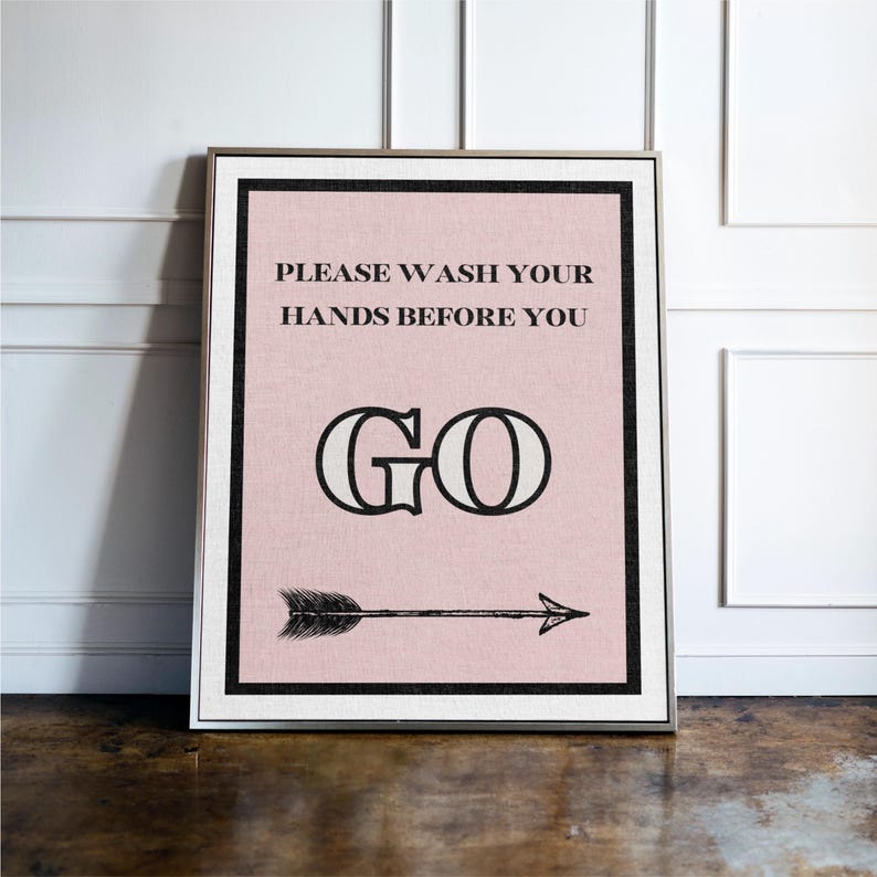 May include: Framed wall art with a pink background and black text that reads "PLEASE WASH YOUR HANDS BEFORE YOU GO." A black arrow points to the right. The frame is silver.