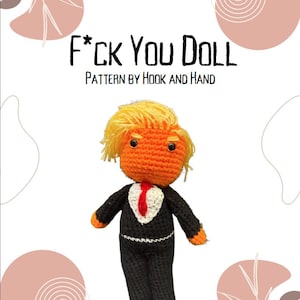 Peut inclure: Poupée au crochet portant un costume noir, une cravate rouge et un visage orange. La poupée a les cheveux blonds et est stylisée pour ressembler à un personnage politique célèbre. Le texte "F*CK YOU DOLL" et "PATTERN BY HOOK AND HAND" est affiché au-dessus de la poupée.