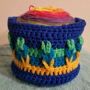 May include: A blue crocheted yarn bowl with a rainbow yarn ball inside. The bowl has a green, blue, and yellow pattern.