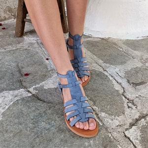 Puede incluir: Un par de sandalias tipo gladiador de color azul con múltiples tiras horizontales sobre el pie, una tira central vertical y una tira ajustable en el tobillo con hebilla de metal. Las sandalias tienen una suela plana color canela y acabado mate.