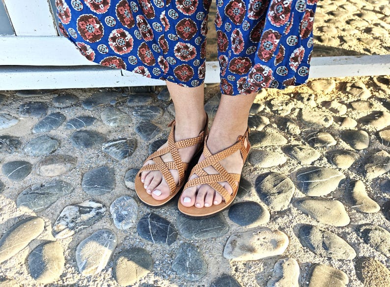 May include: Tan leather sandals with a woven design. The sandals have a crisscross pattern across the top and a buckle closure. The sandals are worn with blue patterned pants. The sandals are on a stone surface.