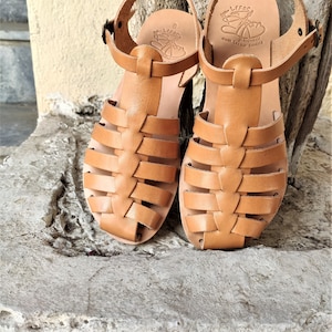 May include: A pair of light brown leather sandals with a woven design on the front. The sandals have ankle straps with buckles and are size 8. The sandals are handmade Greek leather sandals.