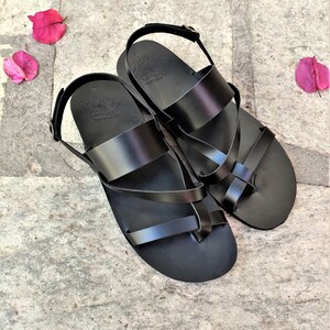 May include: A pair of black leather sandals with a criss-cross strap design. The sandals have a buckle fastening and a toe loop.