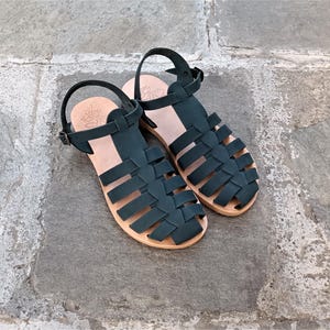 May include: A pair of dark green leather sandals featuring a woven cage design, an adjustable ankle strap with a metal buckle, and a light tan leather insole. The sandals have a flat sole and a closed-toe construction with multiple wide straps.
