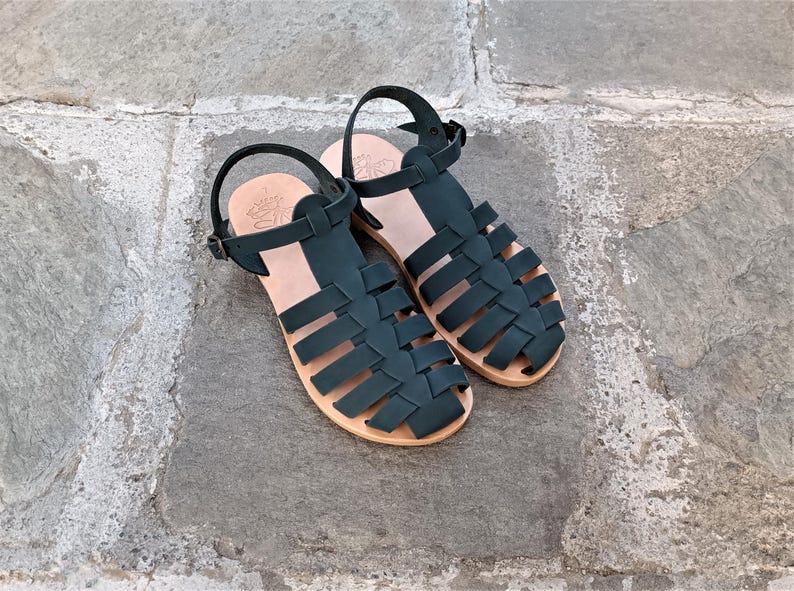 May include: A pair of dark green leather sandals featuring a woven cage design, an adjustable ankle strap with a metal buckle, and a light tan leather insole. The sandals have a flat sole and a closed-toe construction with multiple wide straps.