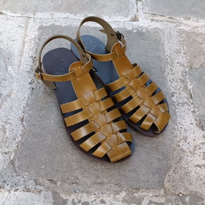 May include: A pair of handcrafted, open-toe sandals with a woven leather design in a warm, golden-brown hue. The sandals feature adjustable ankle straps and rest on a dark sole, perfect for warm weather.