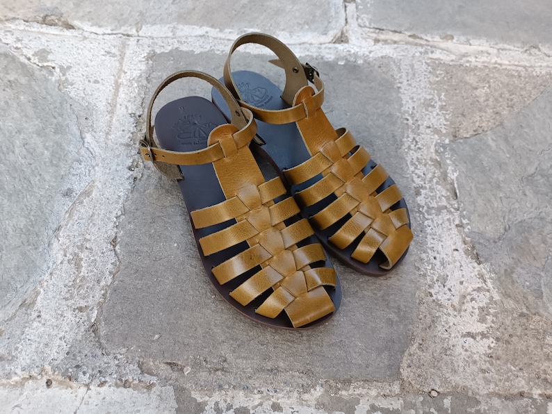 May include: A pair of handcrafted, open-toe sandals with a woven leather design in a warm, golden-brown hue. The sandals feature adjustable ankle straps and rest on a dark sole, perfect for warm weather.