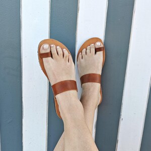 May include: A pair of tan leather sandals featuring a toe loop design and a wide horizontal strap across the midfoot. The sandals have a flat sole and a simple construction, showcasing the natural brown tone of the leather material throughout.