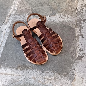 May include: A pair of dark brown leather sandals featuring a woven cage design, an adjustable ankle strap with a gold-tone metal buckle, and a light tan leather footbed with a stamped logo detail on the heel area of each sandal.
