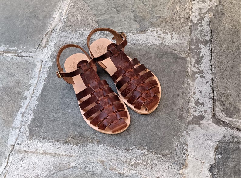 May include: A pair of dark brown leather sandals featuring a woven cage design, an adjustable ankle strap with a gold-tone metal buckle, and a light tan leather footbed with a stamped logo detail on the heel area of each sandal.