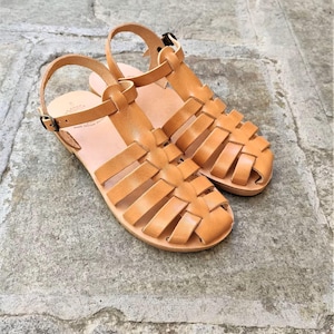 May include: A pair of tan leather fisherman sandals featuring a woven strap design, a rounded toe, and an adjustable ankle strap with a small metal buckle. The sandals have a flat sole and a smooth finish, showcasing a classic cage-style construction.