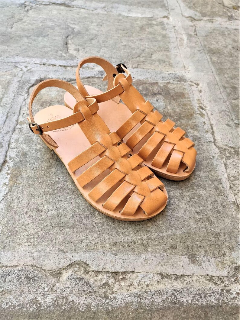 May include: A pair of tan leather fisherman sandals featuring a woven strap design, a rounded toe, and an adjustable ankle strap with a small metal buckle. The sandals have a flat sole and a smooth finish, showcasing a classic cage-style construction.