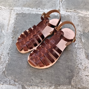 May include: A pair of brown leather sandals with a woven design on the top. The sandals have a light-colored sole and a buckle closure. The sandals are displayed on a stone surface.