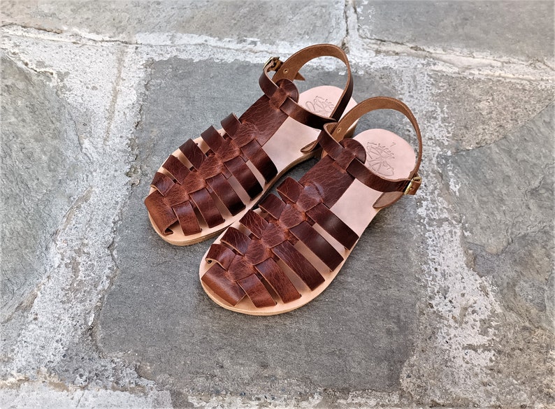 May include: A pair of brown leather sandals with a woven design on the top. The sandals have a light-colored sole and a buckle closure. The sandals are displayed on a stone surface.