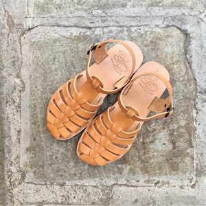 May include: A pair of light brown leather sandals with a woven design on the front. The sandals have a strap around the ankle and a buckle closure. The soles are a lighter shade of brown, and the sandals are sitting on a stone surface.