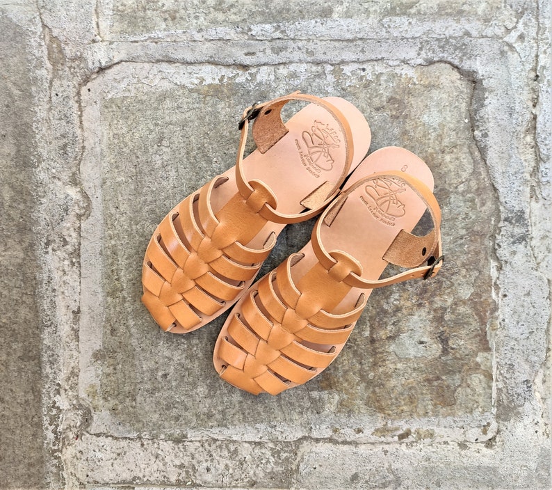 May include: A pair of light brown leather sandals with a woven design on the front. The sandals have a strap around the ankle and a buckle closure. The soles are a lighter shade of brown, and the sandals are sitting on a stone surface.