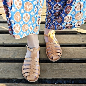 May include: A pair of light brown leather sandals with a cage-like design. The sandals have a strap around the ankle and are set on a wooden surface. The sandals are a summer footwear item.