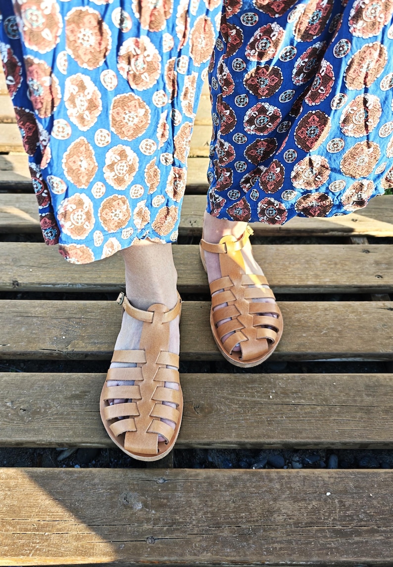 May include: A pair of light brown leather sandals with a cage-like design. The sandals have a strap around the ankle and are set on a wooden surface. The sandals are a summer footwear item.
