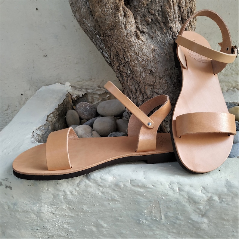 K&ouml;nnte beinhalten: Zwei Paar Sandalen aus braunem Leder mit verstellbaren Kn&ouml;chelriemen. Die Sandalen haben ein schlichtes Design mit einer flachen Sohle.