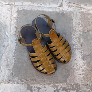 May include: A pair of mustard yellow leather sandals featuring a woven cage design, a closed toe, and an adjustable ankle strap with a metal buckle. The footbed is dark brown with a visible embossed logo stamp near the heel area of each sandal.